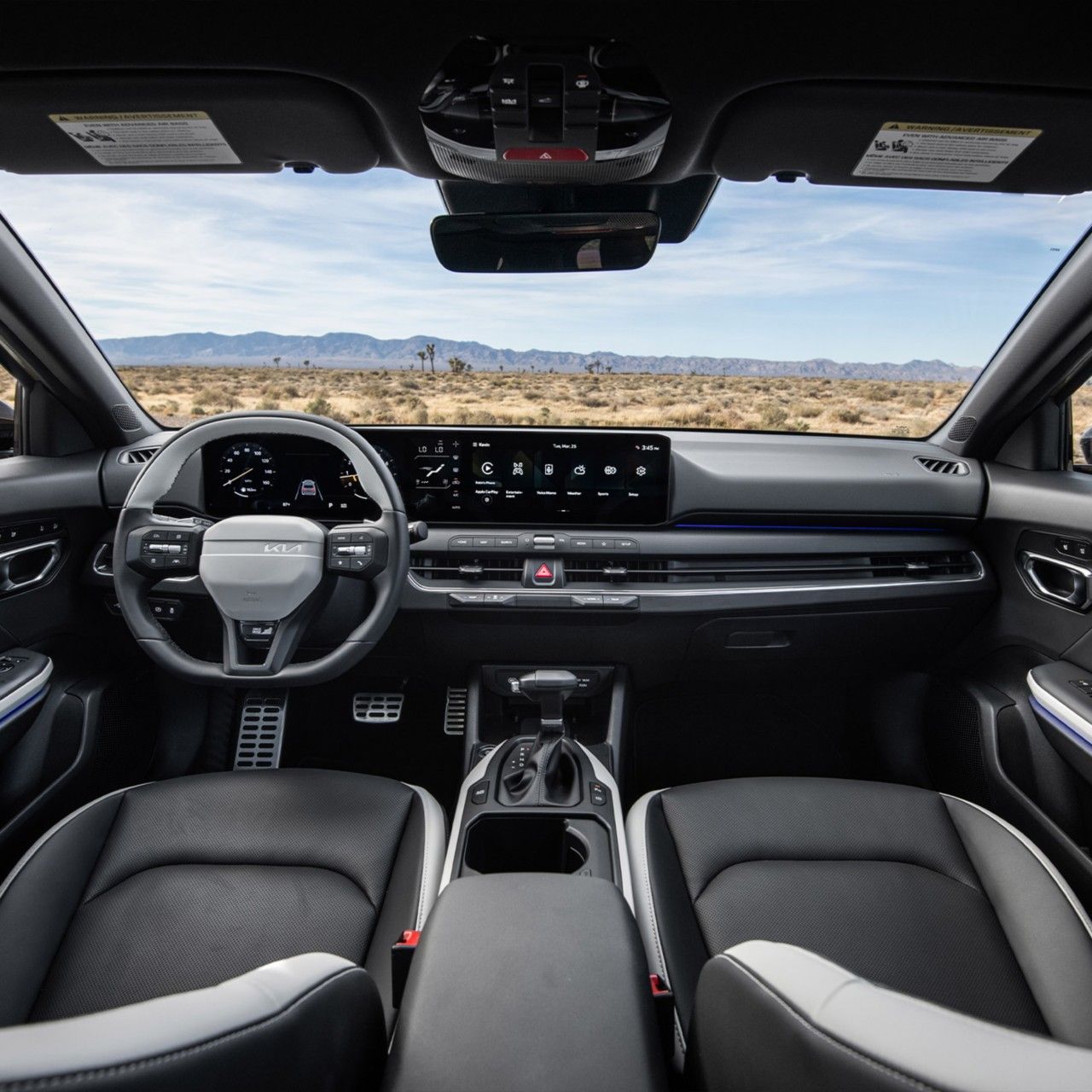  Vista interior completa de los asientos delanteros, la pantalla digital y el volante del Kia K4 Hatchback 2026, con el terreno del desierto visible a través del parabrisas.