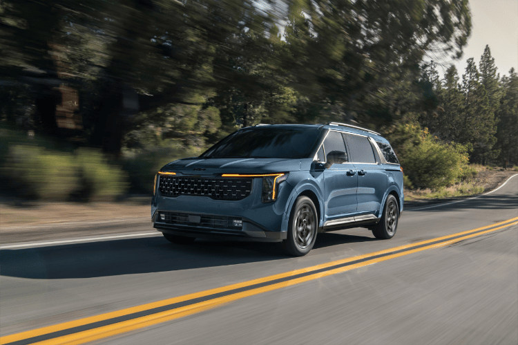 2025 Kia Carnival in blue, driving fast down an empty road near the forest, front three-quarter view