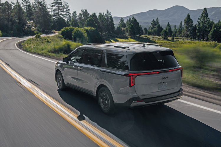 2025 Kia Carnival in gray, driving fast down a curving road near the forest